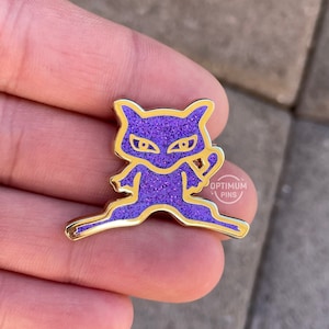 Peut inclure: Épingle en émail violet pailleté avec un contour doré, représentant un personnage stylisé. L'épingle a une bordure dorée et le texte "OPTIMUM PINS" est visible. Un objet de collection.