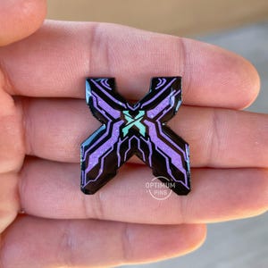 May include: A black and purple enamel pin in the shape of the letter X. The pin has a geometric design with a teal X in the center. The pin is held in a hand. The text "OPTIMUM PINS" is at the bottom.