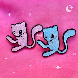 Op de afbeelding: Twee glitter emaille pins met een cartoonachtig katachtig wezen in roze en blauw. De pins hebben zwarte contouren en zijn geplaatst tegen een roze achtergrond met ster- en maangraphics. De tekst "OPTIMUM PINS" is zichtbaar.