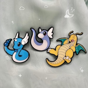 May include: Three enamel pins featuring cartoon characters from the Pokemon franchise. The pins are blue, yellow, and purple. The blue pin is of a blue water-type Pokemon with a blue and white swirl design. The yellow pin is of a yellow dragon-type Pokemon with a green and yellow swirl design. The purple pin is of a purple dragon-type Pokemon with a white and purple swirl design. The pins are all on a light blue background.