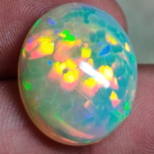 May include: A smooth, oval-shaped cabochon opal gemstone featuring a translucent, milky white base with vibrant flashes of neon yellow, bright orange, and electric blue play-of-color throughout the surface of the polished stone.