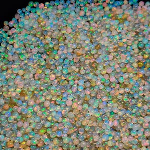 Peut inclure: Une grande collection de petites perles d'opale rondes et translucides. Chaque perle présente une base blanc laiteux avec des éclats irisés de lumière bleue, verte, rose et jaune se reflétant à l'intérieur des surfaces sphériques polies.