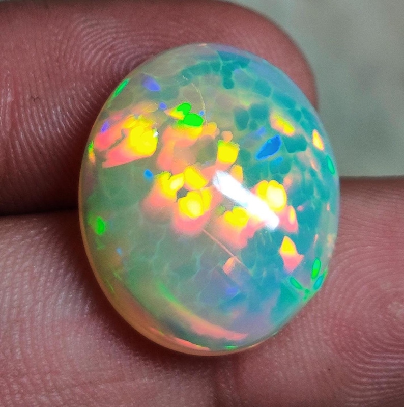May include: A smooth, oval-shaped cabochon opal gemstone featuring a translucent, milky white base with vibrant flashes of neon yellow, bright orange, and electric blue play-of-color throughout the surface of the polished stone.
