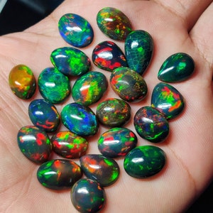 Op de afbeelding: Een verzameling gepolijste zwarte opaal edelstenen met levendige groene, rode en blauwe flitsen. De opalen, in ovale en druppelvormige vormen, worden in een hand gehouden en tonen hun iriserende kleuren. Ideaal voor sieraden.