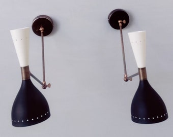 Op de afbeelding: Set van twee wandkandelaars in mid-century modern stijl. Elke wandkandelaar heeft een zwarte, kegelvormige kap met een witte binnenkant en een witte, uitlopende bovenkant. De wandkandelaars hebben messing accenten en zijn op een muur gemonteerd met een donkerbruine cirkelvormige basis.
