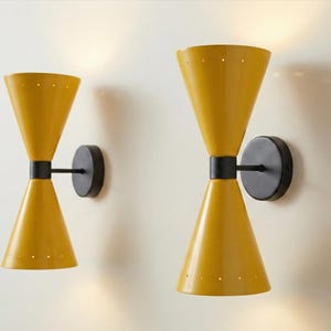 Op de afbeelding: Twee gele wandkandelaars in zandlopervorm met zwarte accenten. Elke kandelaar heeft een ronde basis en een zwarte arm. De kandelaars zijn aan een lichte muur bevestigd en zorgen voor sfeerverlichting.