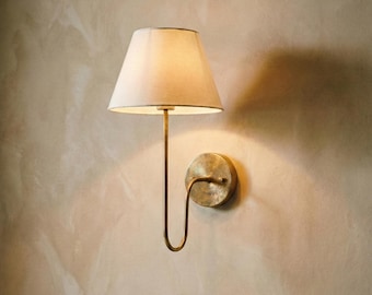 May include: A wall-mounted sconce featuring a cream lampshade and a brass-toned metal frame. The sconce has a circular base and a curved arm supporting the lampshade. The lampshade is a classic cone shape.