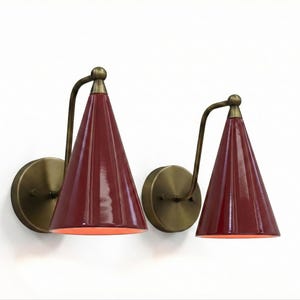 May include: Two wall sconces featuring burgundy cone-shaped shades and brass-toned hardware. Each sconce has a curved arm connecting the shade to a circular wall mount. The shades are a glossy, deep red colour.