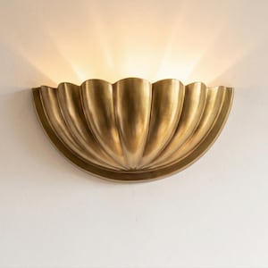 May include: A brass-colored wall sconce with a scalloped design, emitting a warm light. The fixture is mounted on a white wall, creating a contrast. The sconce's shape resembles a stylized shell, adding a touch of elegance to the space.