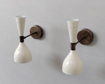Op de afbeelding: Een paar wandlampen met gebroken witte, zandlopervormige kappen met kleine geperforeerde stippenpatronen. Elke lamp is aan de muur bevestigd met een ronde, donkerbronskleurige metalen voet en een bijpassende dunne metalen arm.