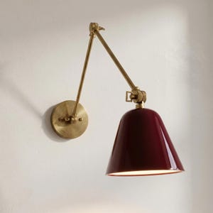 May include: A wall-mounted lamp with a burgundy conical shade and adjustable brass arm. The lamp features a circular base and a pivoting joint for directional lighting. The design is a blend of vintage and modern styles.