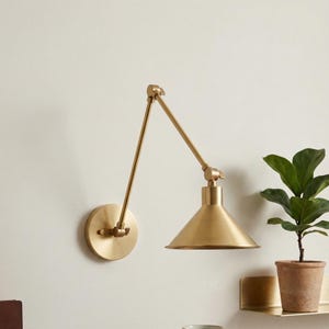 May include: A brass wall lamp with an adjustable arm and a conical shade. The lamp is mounted on a round base and has a warm, golden finish. A small potted plant sits nearby.