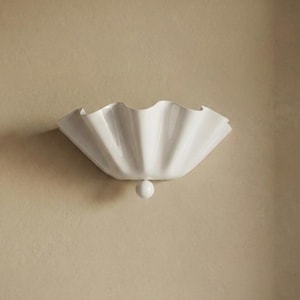May include: A white, shell-shaped wall sconce with a spherical base, mounted on a beige wall. The lighting fixture has a scalloped edge. This decorative piece is designed for interior spaces.