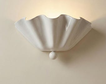 May include: A white, scalloped wall sconce with a glossy finish. The light fixture has a shell-like design and a small, round accent at the bottom. The sconce is mounted on a light beige wall.