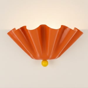 Könnte beinhalten: Orangefarbene Wandleuchte mit einem gerüschten, fächerartigen Design. Die Leuchte hat eine glatte, glänzende Oberfläche und eine kleine gelbe Kugel am unteren Ende. Die Leuchte ist an einer hellen Wand montiert.