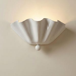Może przedstawiać: Biały ceramiczny kinkiet ścienny o falistym, przypominającym muszlę kształcie. Oprawa oświetleniowa ma zaokrągloną podstawę i mały, kulisty detal. Kinkiet jest zamontowany na jasnobeżowej ścianie.