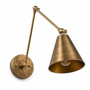 Può includere: Una lampada da parete a braccio oscillante con finitura in ottone anticato. L'apparecchio presenta una piastra circolare a muro, due bracci metallici regolabili collegati da uno snodo e un paralume conico con una lampadina a filamento.