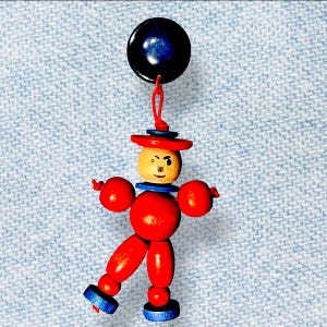 May include: A whimsical wooden figure with a red body, arms, and legs, a tan head, and a red hat. The figure has blue accents and is suspended from a dark blue button with a red string. The figure is a decorative item.