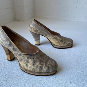 Può includere: Un paio di tacchi alti beige con plateau e motivo a pelle di serpente. I tacchi sono alti circa 10 cm e hanno una piattaforma spessa.