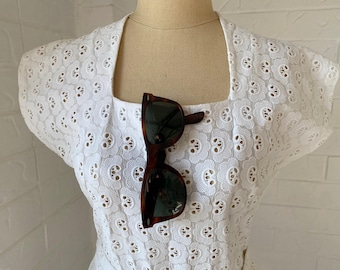 May include: White eyelet dress with a square neckline and short sleeves. A pair of brown sunglasses are resting on the dress.