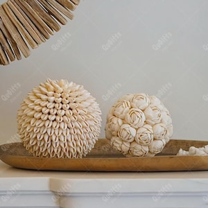 Könnte beinhalten: Zwei weiße Dekorationssphären aus Muscheln auf einem goldenen Tablett. Die Kugel links besteht aus kleinen Muscheln und die Kugel rechts aus größeren Muscheln, die in Blumenform angeordnet sind.