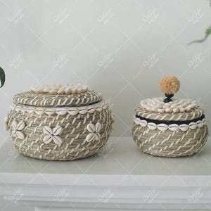 Può includere: Due cesti portaoggetti intrecciati con coperchi. Il cesto più grande presenta un motivo floreale realizzato con conchiglie bianche. Entrambi i cesti sono adornati con conchiglie attorno ai bordi. I coperchi sono sormontati da conchiglie e un pompon decorativo.