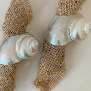May include: Two seashell napkin rings with an iridescent, pearlescent finish. Each ring is wrapped around a burlap napkin. The shells have a spiral design. The Etsy shop name is visible. The background is neutral.