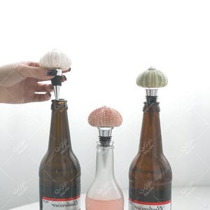 May include: Three decorative bottle stoppers with sea urchin designs. One is being inserted into a brown bottle. The stoppers have white, pink, and green tops with silver and black bases. The bottles are various shades of brown and clear.