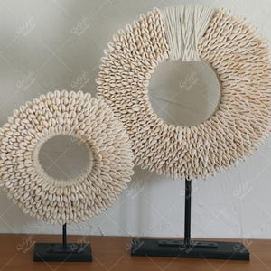 Könnte beinhalten: Zwei dekorative Muschelkranz-Skulpturen auf schwarzen Metallständern. Die Kränze sind kreisförmig, mit einem Ring aus Muscheln um eine zentrale Öffnung. Der größere Kranz hat oben einen Abschnitt aus weißem Seil.