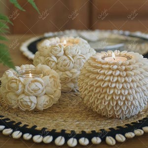 May include: Three cream-colored seashell candle holders with lit candles inside. The candle holders are arranged on a woven mat with a black border. The candle holders are different sizes and shapes, with the largest one having a flower-like design.