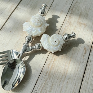May include: A silver-toned metal spoon and fork set featuring handles adorned with iridescent white seashell accents. The handles transition from a polished metallic base to a detailed, spiraled shell finial, creating a distinct decorative look.