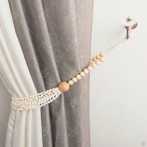 Può includere: Un fermatenda decorativo con una fila di piccole conchiglie bianche e una grande perla di legno. Il fermatenda tiene indietro una tenda grigia. Un gancio di cristallo trasparente è montato a parete.