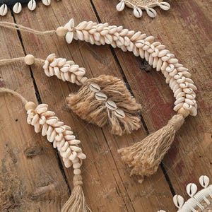 May include: Decorative seashell garlands and tassels. The garlands are made of small, cream-coloured seashells strung together with natural twine. Tassels are made of natural fibre with seashells attached. The items are arranged on a rustic wooden surface.