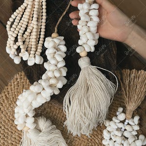 May include: Decorative seashell and wooden bead garlands. The garlands feature white seashells, wooden beads, and natural fiber tassels. The items are displayed on a wooden surface and a woven mat.