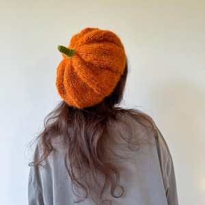 May include: A knitted beret in a vibrant orange pumpkin shape, featuring a textured, fuzzy yarn construction and a small, pointed green stem detail at the center top, designed to be worn on the head with a rounded, voluminous silhouette.