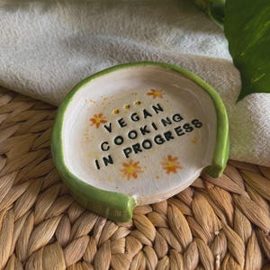 May include: A ceramic spoon rest with a green rim and the text "VEGAN COOKING IN PROGRESS". The spoon rest is off-white with orange floral accents. The spoon rest is round with a small lip.
