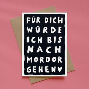Könnte beinhalten: Eine rechteckige Grußkarte mit dunkelblauem Hintergrund und weißem, handgeschriebenem Text, der FÜR DICH WÜRDE ICH BIS NACH MORDOR GEHEN lautet, mit einem kleinen weißen Herzsymbol am Ende, platziert auf einem hellbraunen Papierumschlag.