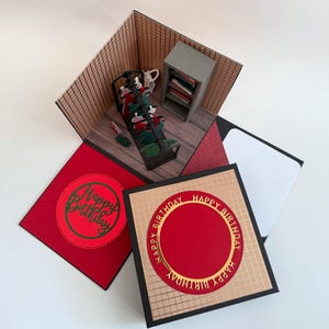 Könnte beinhalten: Eine handgefertigte Pop-up-Geburtstagskarte mit einer Miniatur-Raumszene. Die Karte hat ein rot-goldenes Farbschema mit den Worten "Happy Birthday" auf der Vorderseite. Im Inneren ist ein kleiner Raum mit Möbeln zu sehen.