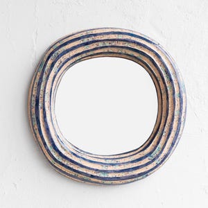 Circular decorative ceramic mirror with a cream-coloured frame featuring blue and white stripes. The mirror is centred against a white wall. The frame has a textured, ridged design, adding visual interest to the piece.
