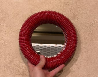 May include: A circular mirror featuring a glossy, deep red frame with a distinct ribbed or corrugated texture. The reflective glass center is surrounded by the thick, rounded, and textured red border, creating a bold contrast.