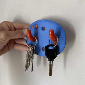 Op de afbeelding: Een blauwe ovale wand-sleutelhouder van mat materiaal met twee feloranje gebogen haken. De houder heeft twee ronde gaten voor montage en twee kleine oranje decoratieve stippen. Twee metalen sleutelringen met sleutels zijn bevestigd.