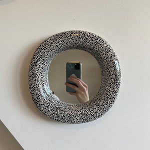 以下が含まれることがあります： 黒い水玉模様の白い枠が付いた丸い鏡。鏡にはスマートフォンを持った人が映っている。