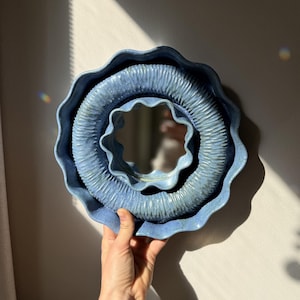 Può includere: Uno specchio in ceramica blu con un design unico a strati. Lo specchio ha una cornice circolare ondulata con dettagli strutturati. La cornice è di colore azzurro e lo specchio stesso è visibile al centro. Lo specchio è tenuto contro un muro neutro.