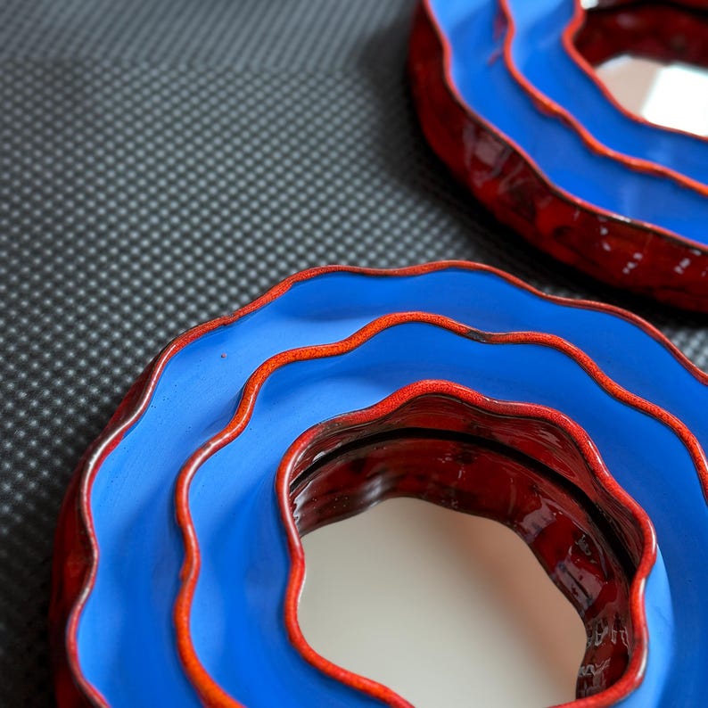 Pu&ograve; includere: Due ciotole decorative in ceramica con un design ondulato. Le ciotole hanno un esterno blu brillante con bordo e interno rossi. Le ciotole sono posizionate su una superficie grigio scuro.