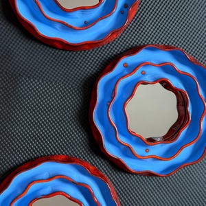 Pu&ograve; includere: Tre specchi decorativi dal design unico. Ogni specchio presenta una cornice circolare e ondulata con un interno blu vibrante e un bordo rosso a contrasto. Gli specchi hanno una superficie riflettente.