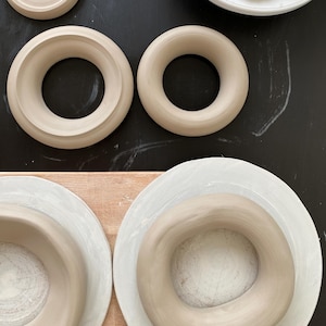 Pu&ograve; includere: Quattro anelli in ceramica di diverse dimensioni, con una glassa marrone chiaro, sono disposti su una superficie nera e di legno. Gli anelli sono tutti circolari e hanno un foro al centro.