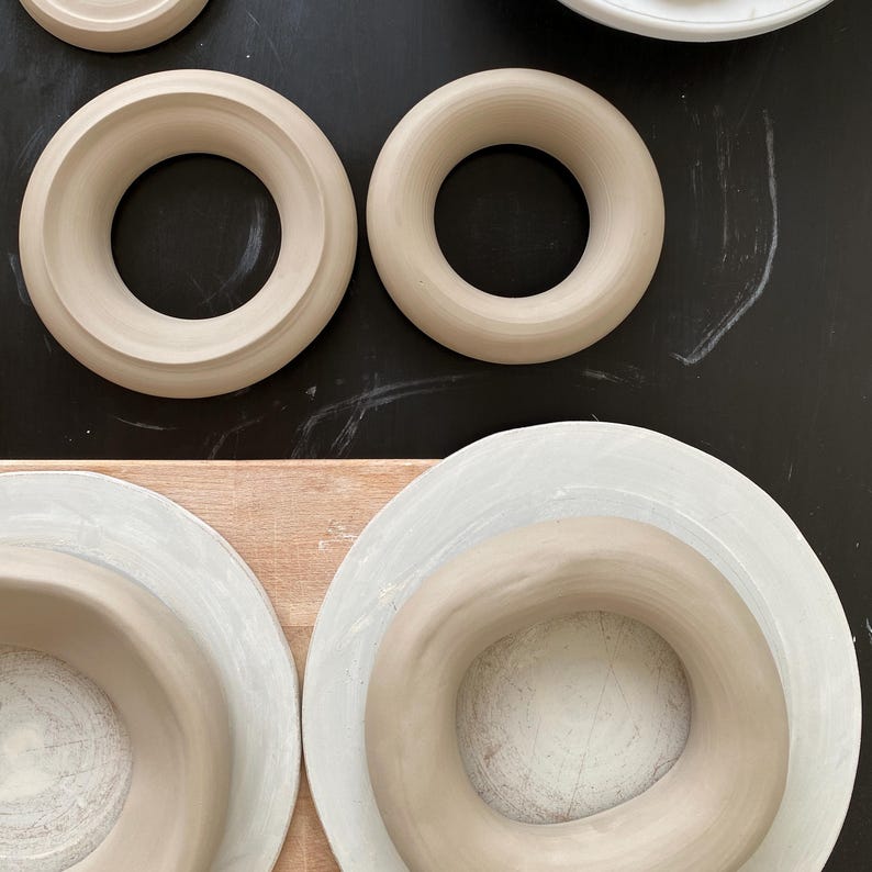Pu&ograve; includere: Quattro anelli in ceramica di diverse dimensioni, con una glassa marrone chiaro, sono disposti su una superficie nera e di legno. Gli anelli sono tutti circolari e hanno un foro al centro.