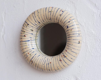 Pode incluir: Um espelho redondo com uma moldura creme. A moldura é decorada com riscas azuis verticais e salpicos coloridos. O espelho está montado numa parede branca.
