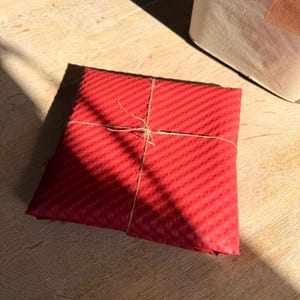 Op de afbeelding: Een vierkant cadeau verpakt in rood, getextureerd papier en vastgebonden met touw. Het papier heeft een herhalend patroon. Het pakket ligt op een licht houten oppervlak.