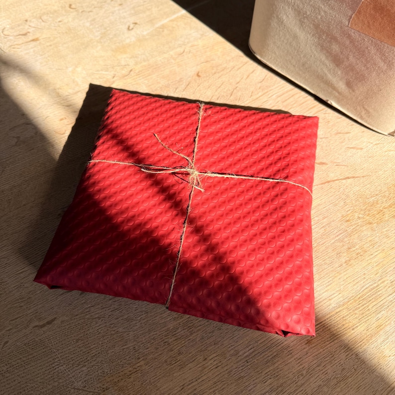 Op de afbeelding: Een vierkant cadeau verpakt in rood, getextureerd papier en vastgebonden met touw. Het papier heeft een herhalend patroon. Het pakket ligt op een licht houten oppervlak.